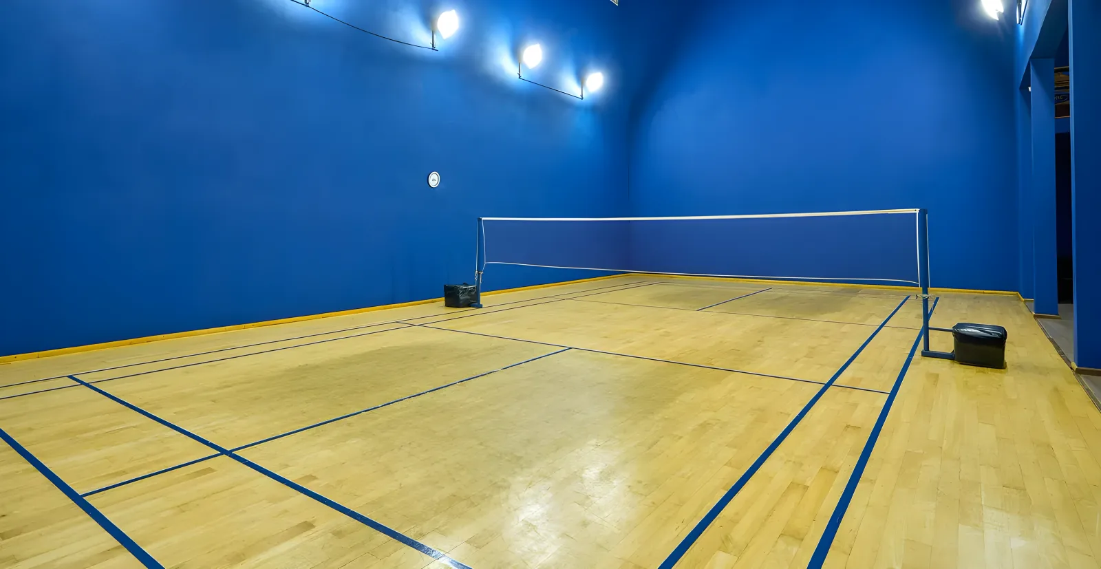 Empty pale wooden badminton court at Colombo Swimming Club, net up between deep blue walls, with blue court markings and wall-mounted floodlights